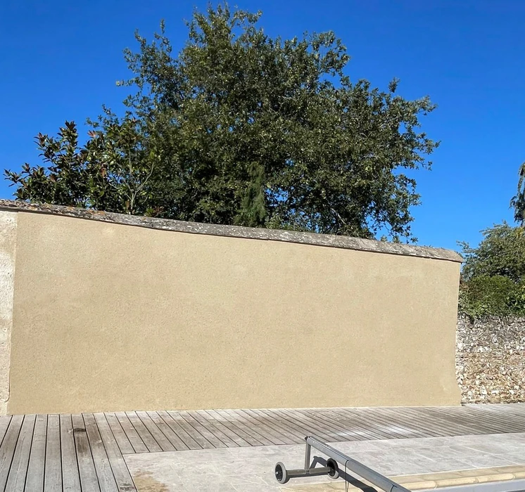 Mur extérieur en béton ou crépi beige avec couronnement en pierre naturelle, situé dans un environnement paysager avec végétation mature